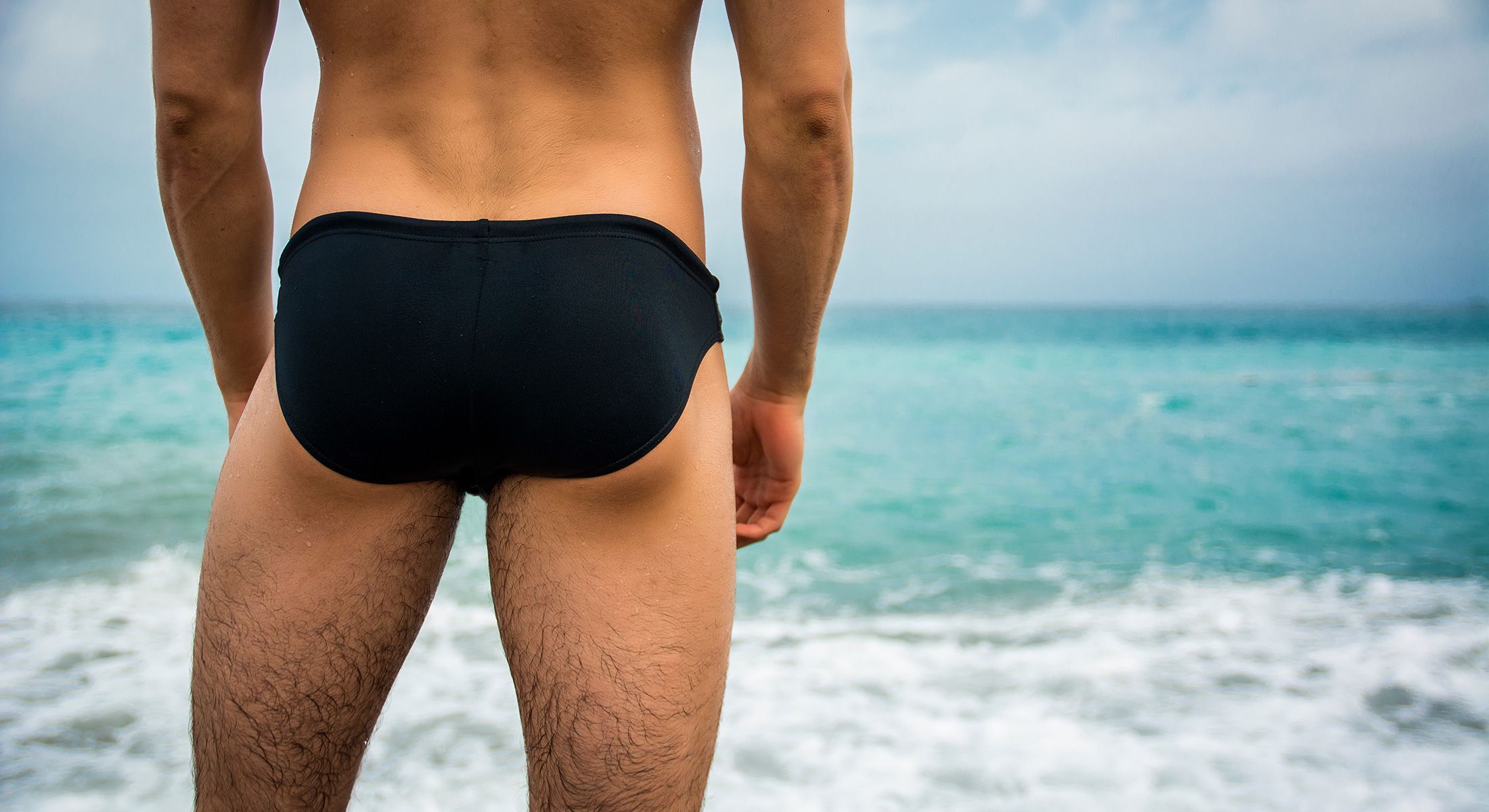 Man standing at beach in swimwear looking at the sea, lower body crop